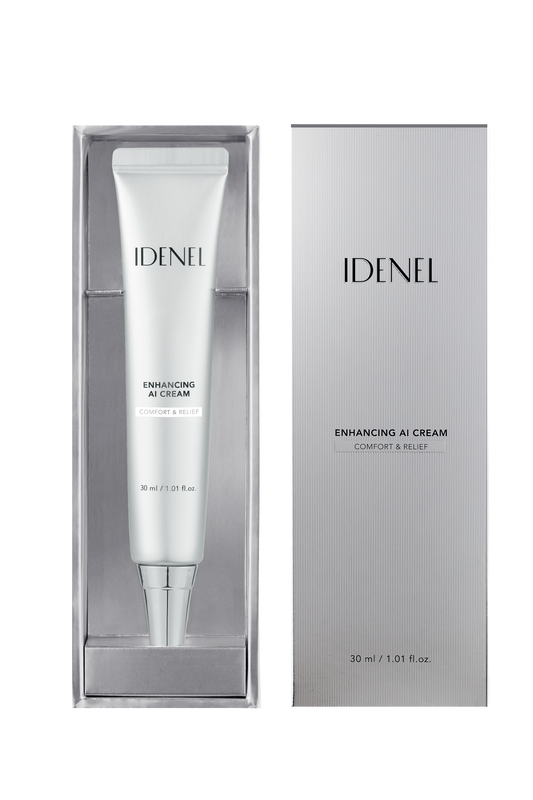 A white cosmetic cream tube labeled IDENEL ENHANCING AI CREAM sits inside a clear rectangular display box, with the matching silver-gray product box standing beside it on a white background.